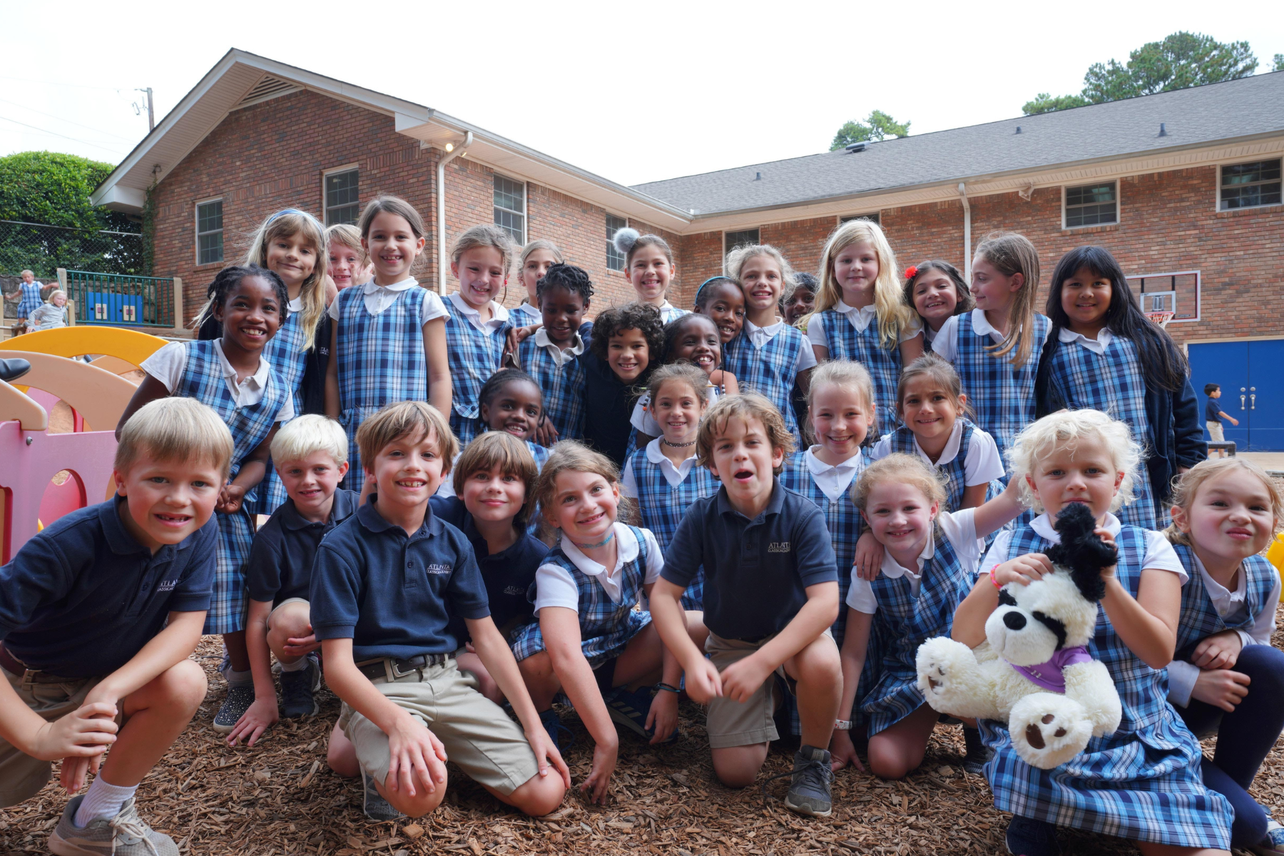 Large group of smiling elementary school students wearing plaid school uniforms posing together on a playground with a brick school building in the background. The children are gathered closely, radiating joy and energy, while one student holds a plush panda toy, creating a cheerful and inclusive outdoor scene.