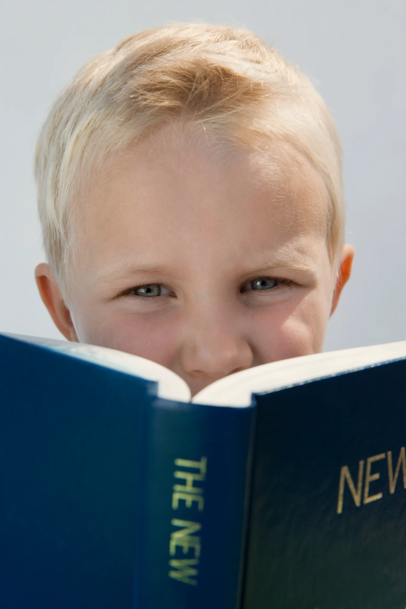 A young boy with short blonde hair and bright blue eyes peers over the top of a large blue book titled 'The New.' He has a curious and focused expression, suggesting deep engagement in reading. The background is a clean, neutral white, drawing attention to his face and the book.