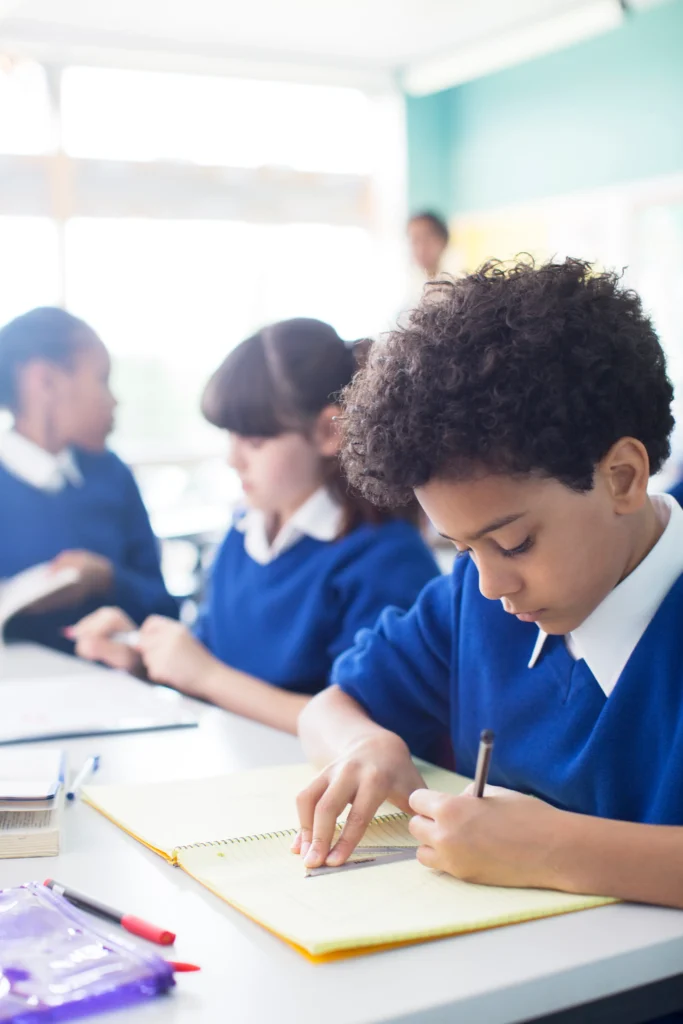 A young student in a blue school uniform is deeply focused on drawing with a ruler in a notebook, surrounded by classmates engaged in their studies. The bright and modern classroom setting reflects an atmosphere of concentration and learning.