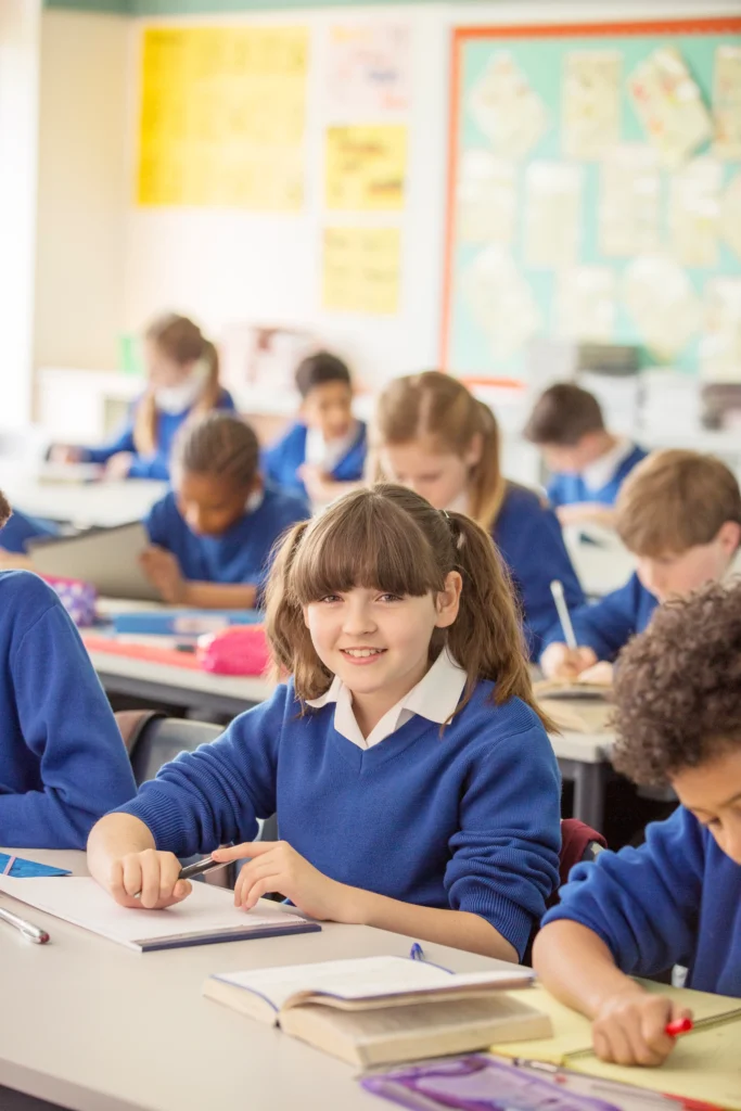 A cheerful young girl in a blue school uniform sits at her desk in a bright and lively classroom, surrounded by classmates engaged in their studies. She smiles at the camera while holding a pen, with an open book and stationery on her desk, reflecting an atmosphere of active learning.