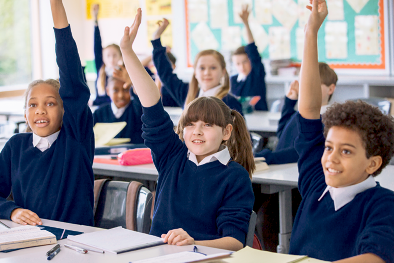 Children in a classroom raising their hands enthusiastically, dressed in matching navy-blue uniforms, seated at desks with open notebooks and pens, showcasing an engaged and active learning environment.