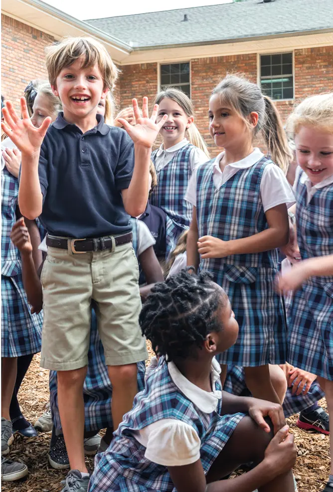 Group of smiling schoolchildren outdoors on a playground, with a boy in the foreground enthusiastically raising his hands. The students are wearing plaid school uniforms and enjoying a lively moment of interaction, with a brick school building in the background.