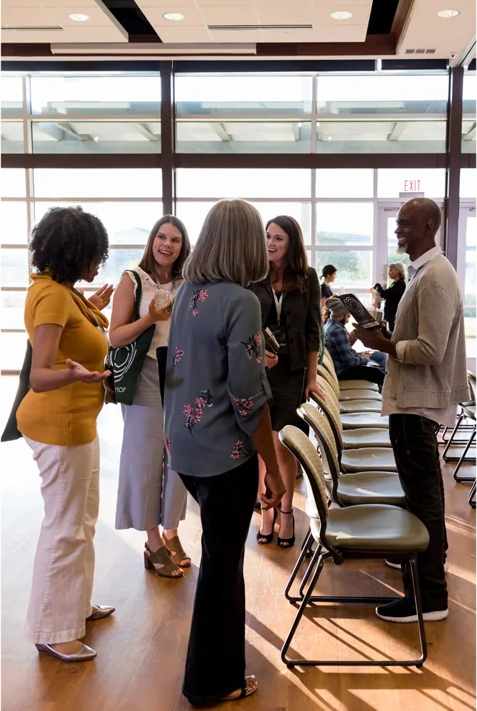 Group of professionals engaged in a friendly conversation in a bright, modern conference room with large windows. The individuals are standing near a row of chairs, smiling and interacting, creating a welcoming and collaborative atmosphere at a networking or business event.